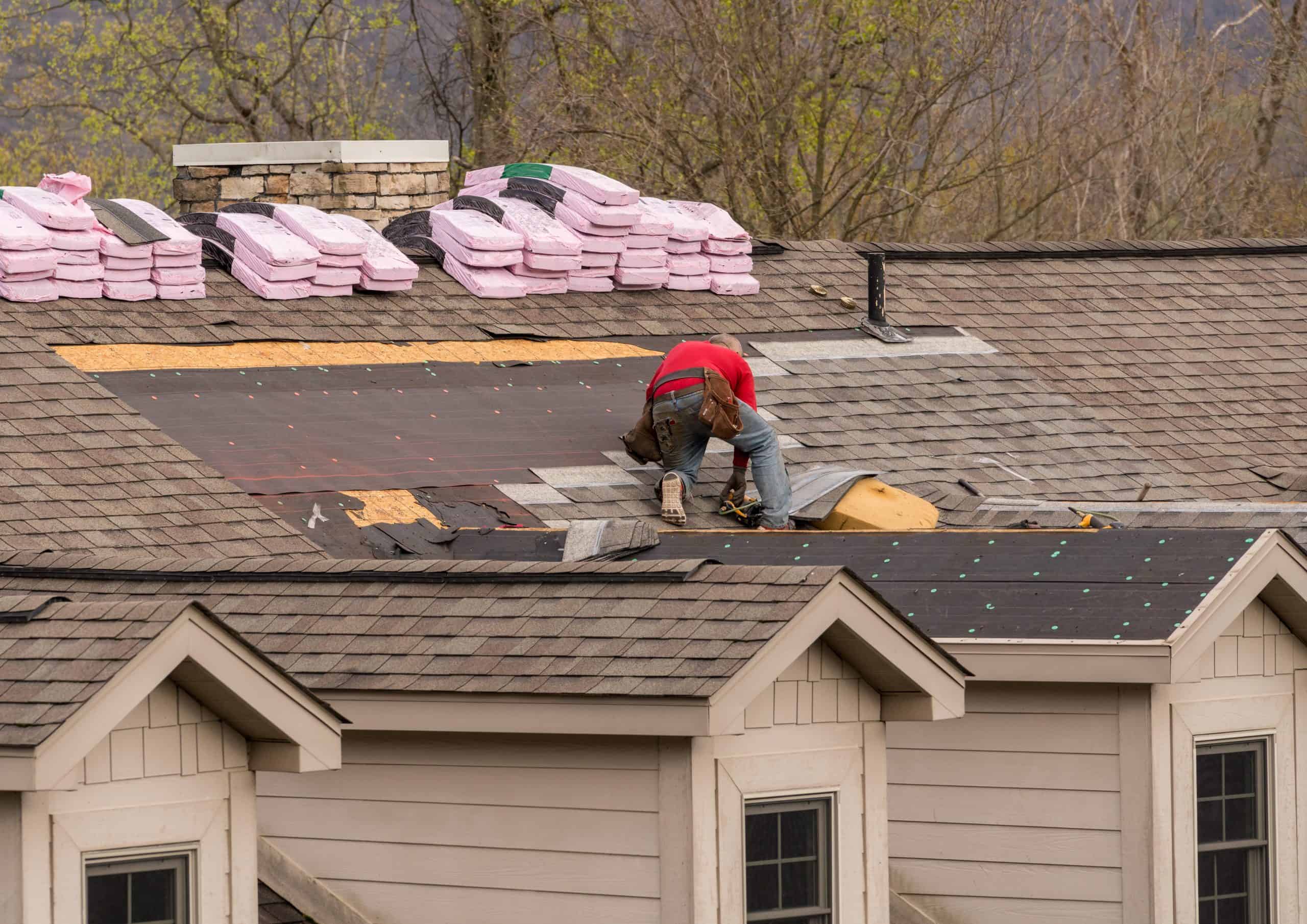 A construction worker in a red shirt diligently handles a shingle rejuvenation on a house roof in Franklin County, OH. Bundles of pink insulation are stacked nearby, while trees and building details provide a scenic backdrop.