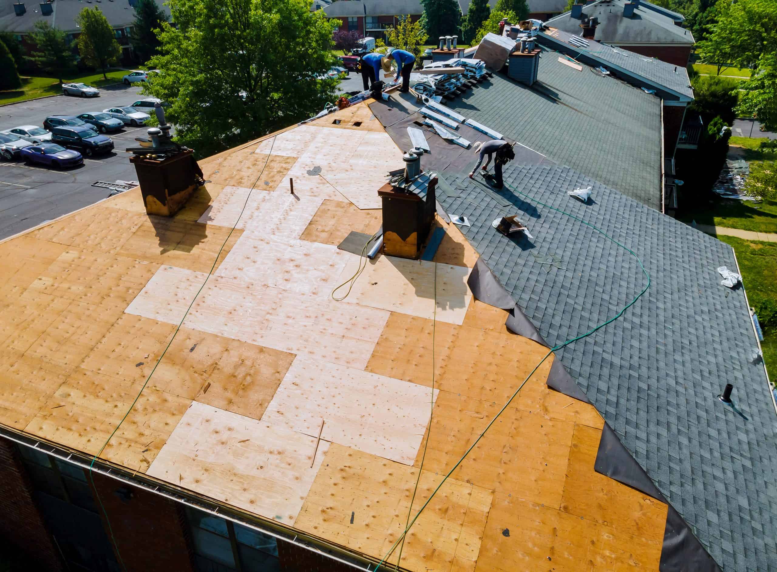Aerial view of a roof under renovation with several construction workers involved in roof renewal in Franklin County OH. Parts of the roof are covered with plywood, others with shingles. Nearby are parked cars, trees, and a street.