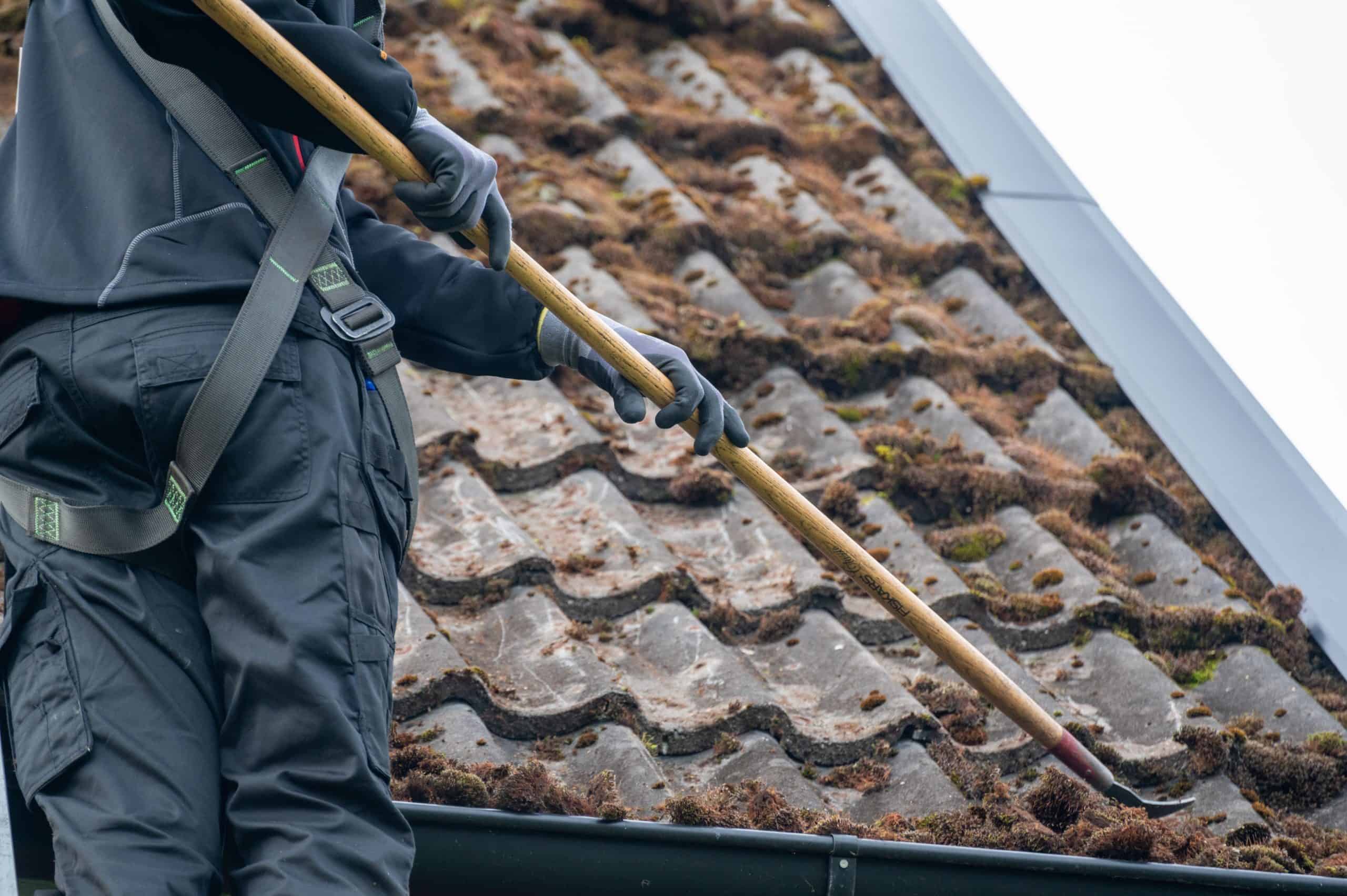 A person in protective gear and gloves uses a long brush to remove moss from a tiled roof under cloudy skies, showcasing professional roof cleaning services in Franklin County, OH.
