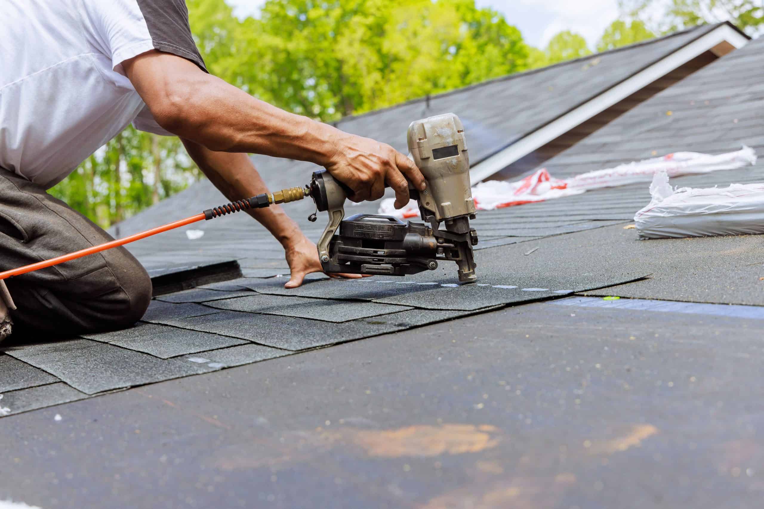 A person in a white shirt uses a nail gun to install asphalt shingles on a roof, part of a Roof Renewal Franklin County OH project. The background shows trees and a clear sky, while roof materials are scattered on the rooftop.